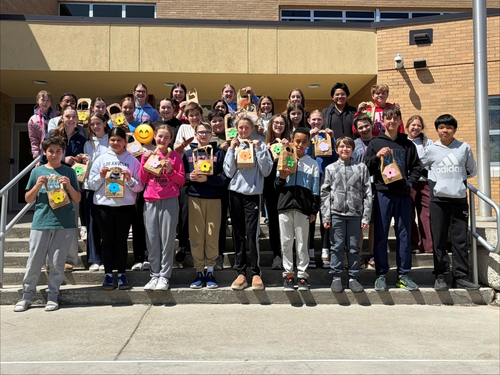 A large group of middle school students stands on the front steps outside a school building on a sunny day, smiling and holding small handmade May Day baskets decorated with colorful paper flowers.