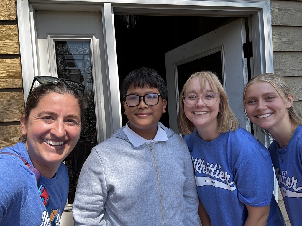 An incoming Whittier Middle School students stands smiling in the doorway of their home next to Whittier Middle School staff members also smiling. 