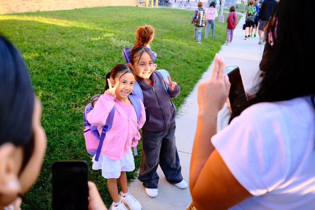 Two elementary students excitedly do the "peace" sign with their fingers while a parent takes their photo on the first day of school 