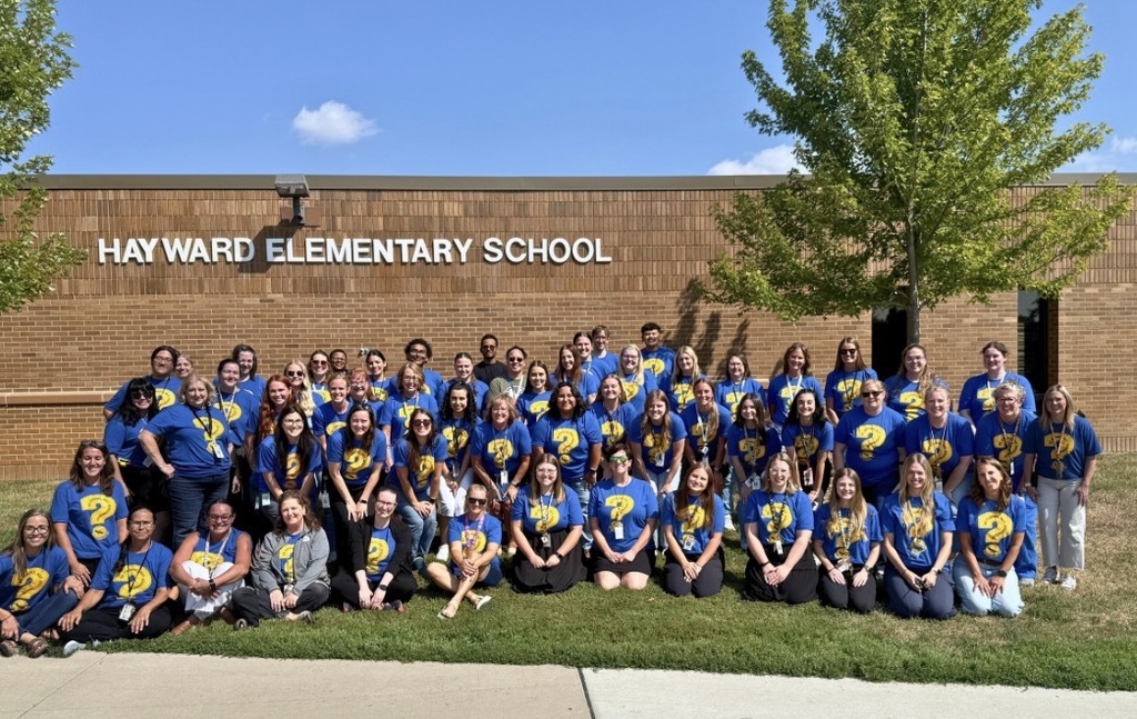 Hayward staff in front of the school building, wearing matching blue shirts that feature a large yellow question mark with the words "be curious, not judgmental"