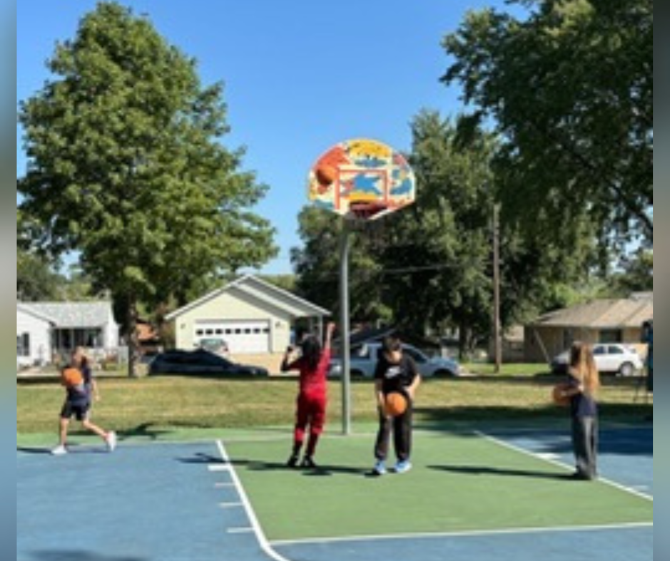 Laura B. Anderson Elementary students throw a basketball towards a basketball hoop with a newly designed artistic backboard at an unveiling celebration at Pioneer Park