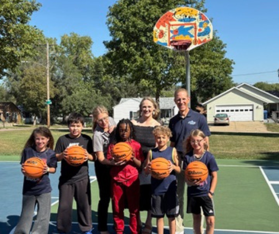 Laura B. Anderson Elementary students stand holding a basketball in front of a basketball hoop that has newly designed backboards at an unveiling ceremony at Pioneer Park