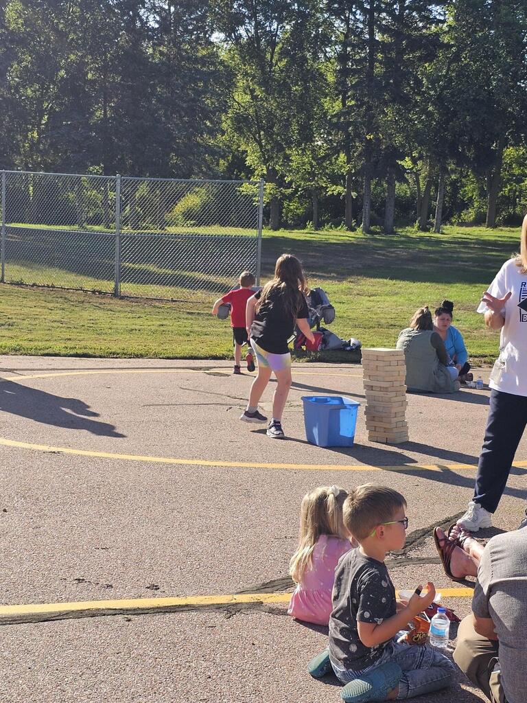 kids sitting on the ground eating and talking, another girl playing large Jenga