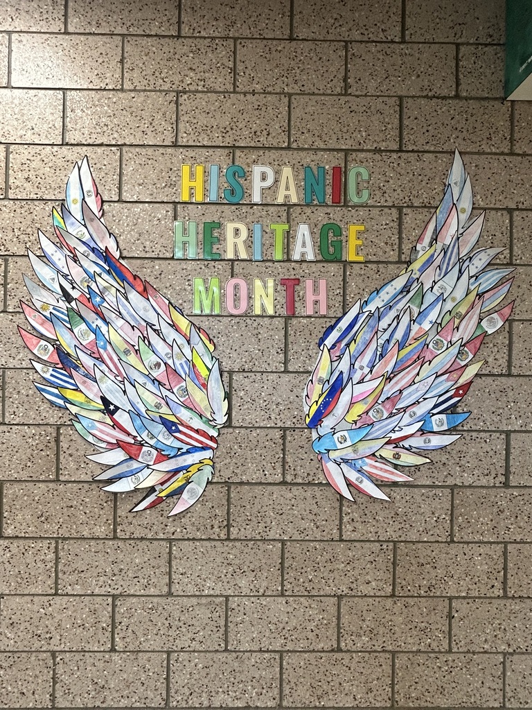 A hallway display at Memorial Middle School showing two large wings made of hand-colored paper feathers, each feather decorated with the flag of a Spanish-speaking country. Colorful cut-out letters between the wings read "Hispanic Heritage Month."