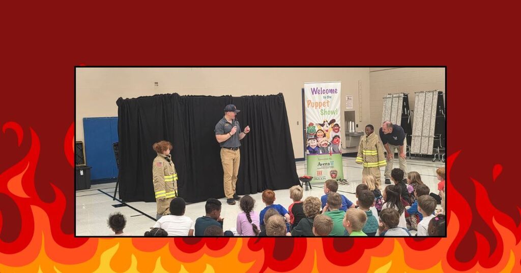 Cartoon fire, students observing puppet show with firefighter presenting, students wearing firefighter gear