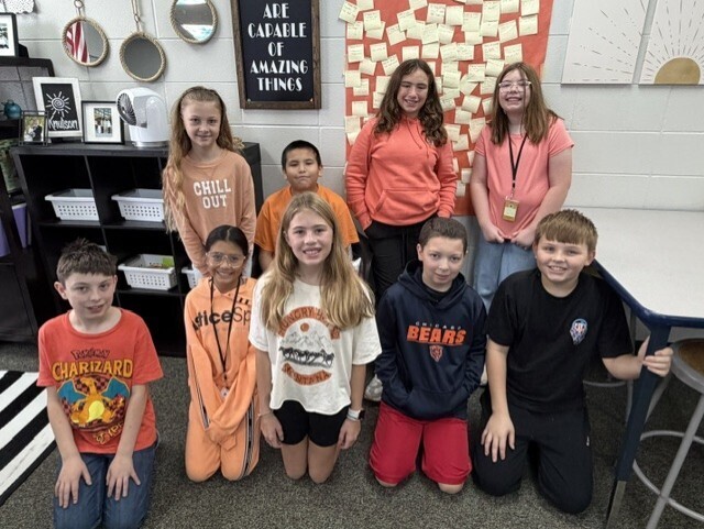 Nine Memorial Middle School students in orange shirts pose together in a classroom. A sign on the wall behind them reads "You are capable of amazing things."