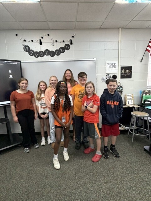 A Memorial Middle School teacher and seven students wearing orange stand together in a classroom beneath a banner reading "Mavericks."