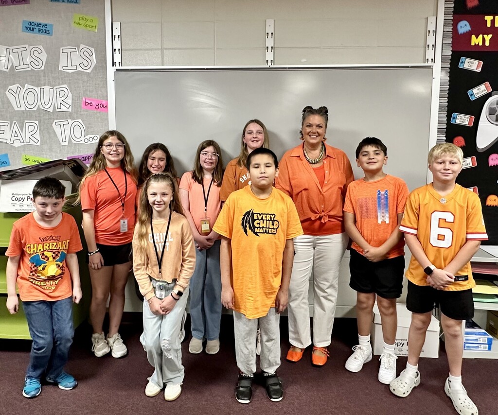 A Memorial Middle School teacher stands with nine students in a classroom, all wearing orange for Orange Shirt Day. One student's shirt reads "Every Child Matters." A bulletin board behind them reads "This is your year to…"