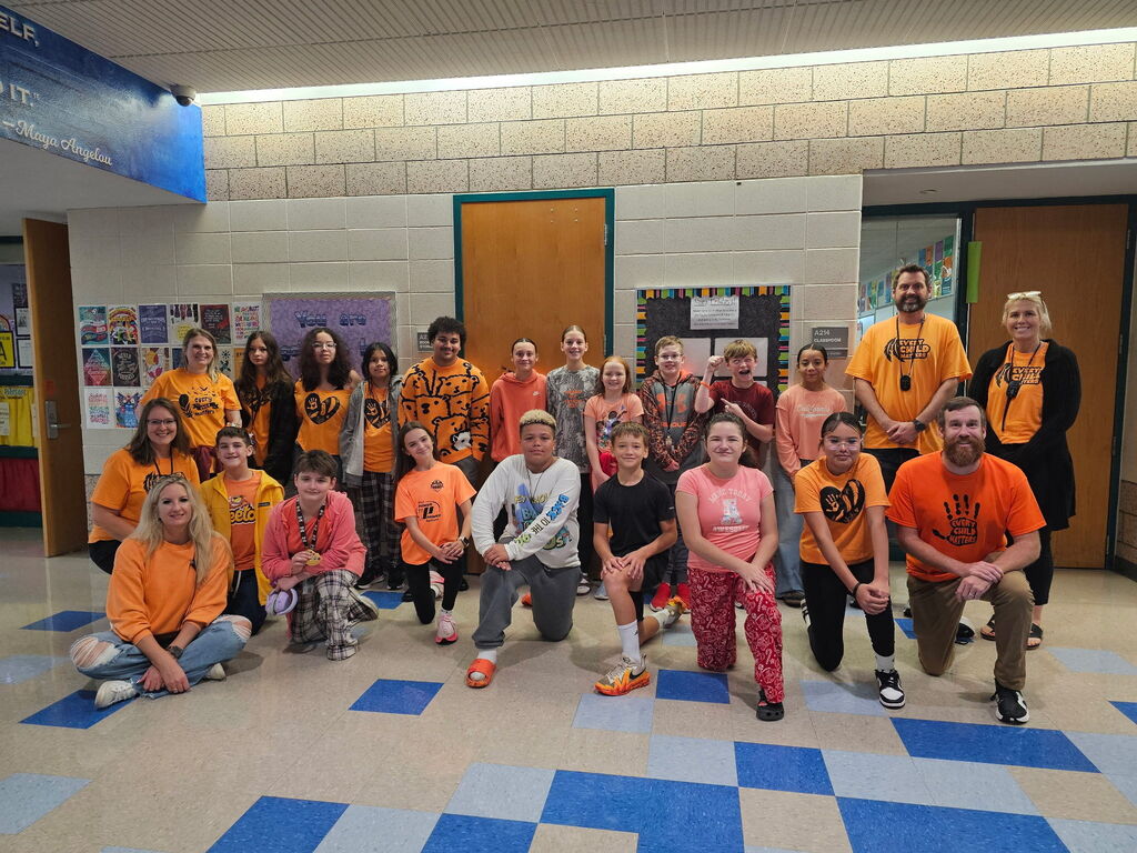 A large group of Memorial Middle School students and staff gathered in a hallway for Orange Shirt Day, all wearing orange. Several shirts read "Every Child Matters" with an orange handprint and feather design.