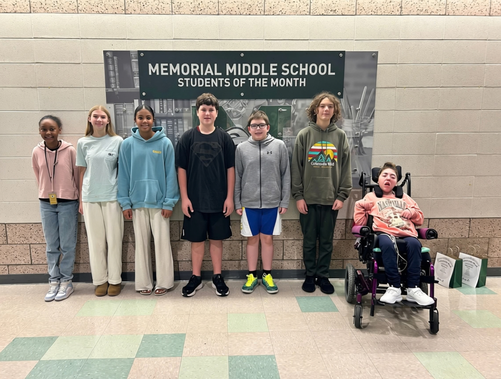 Seven Memorial Middle School students posing together under the "Memorial Middle School Students of the Month" wall sign.