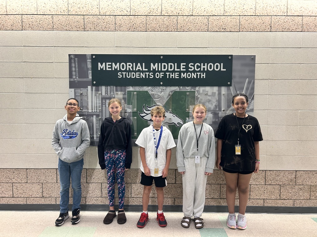 Five Memorial Middle School students smiling in front of the "Memorial Middle School Students of the Month" display wall.