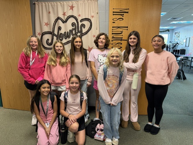 Nine smiling students dressed in pink pose in a school hallway outside Mrs. Knutson’s classroom, in front of a banner that reads “Howdy.”