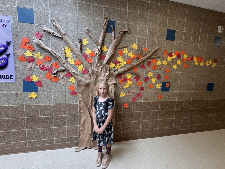 girl standing next to a paper tree sculpture hanging on a wall with fall leaves of yellow red and orange