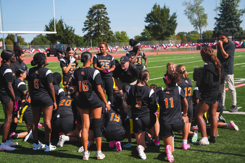 Washington High School Girls Flag Football players stand on the sideline in a huddle listening to their coach at the first Girls Flag Football Jamboree at Howard Wood Field