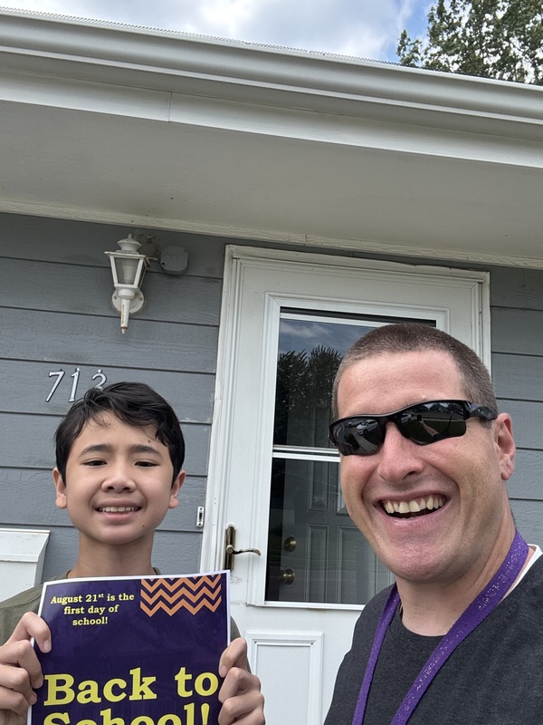 a  middle school student smiles at the camera next to his teacher holding a back to school sign