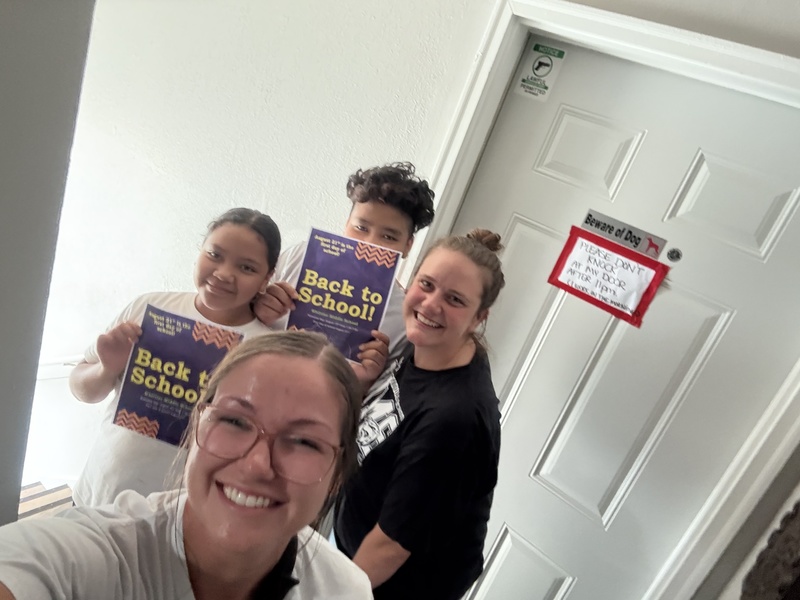 four individuals stand in front of a white door holding back to school signs