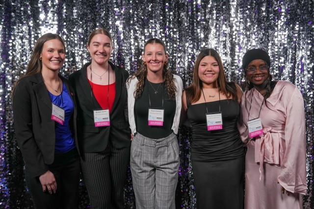 The five scholarship finalist women dressed formally stand with arms around one another in front of a sparkly grey decorative backdrop with excited looks on their faces