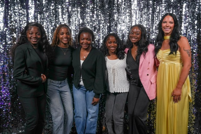 Six women dressed formally stand with arms around one another in front of a sparkly grey decorative backdrop with excited looks on their faces