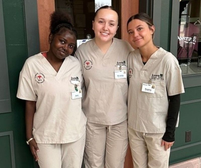 Three students stand smiling with arms around one another in their beige scrubs at Avera Health where they are partaking in an internship