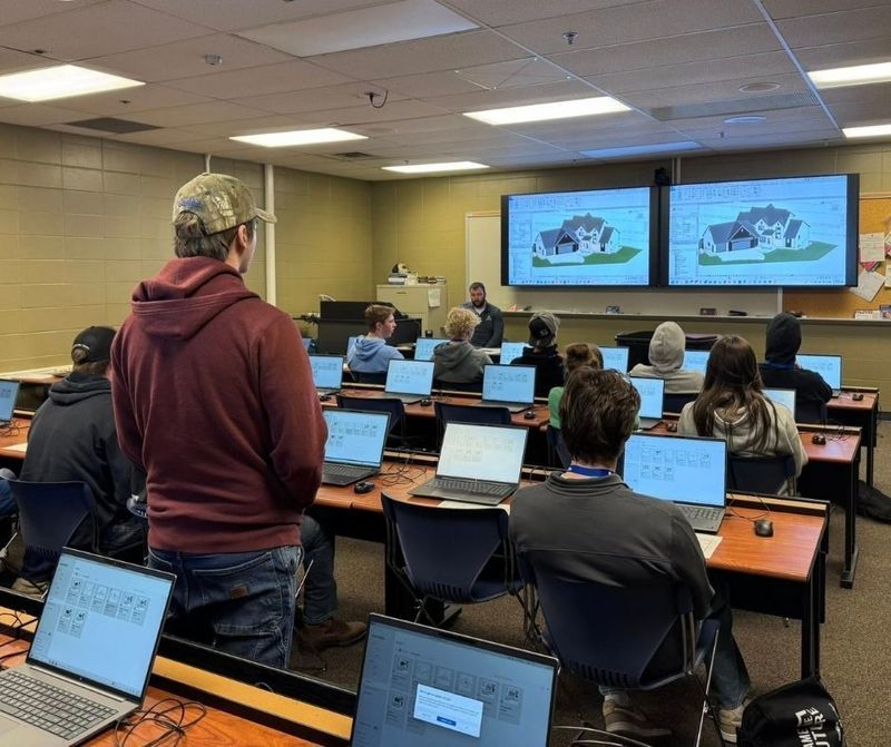 Several high school students fill a classroom with their laptops open but looking straight ahead at a large tv with an image of a house rendering