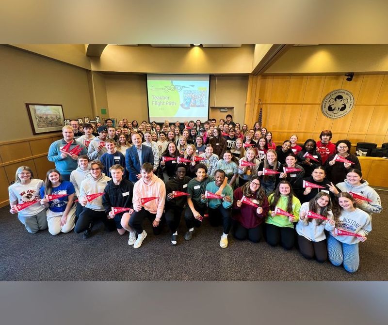 Over 100 teacher pathway students huddle together smiling at the camera holding USD flags
