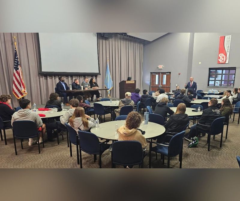 Middle school students sit around sevral circular tables listening to a panel of speakers