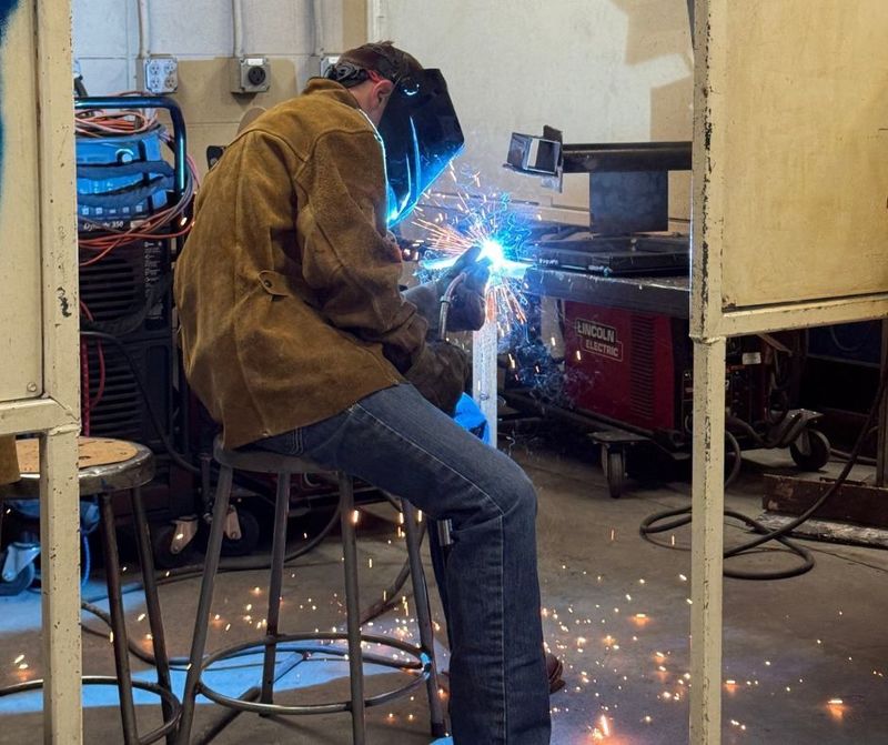 Student sits on a stool with a welding jacket and face protectant on welding an object with blue sparks flying