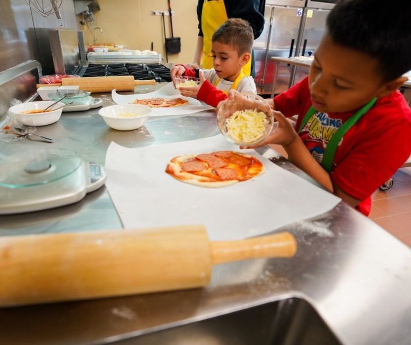 Two elementary students stand in an industrial kitchen at CTE sprinkling cheese on homemade pizza dough