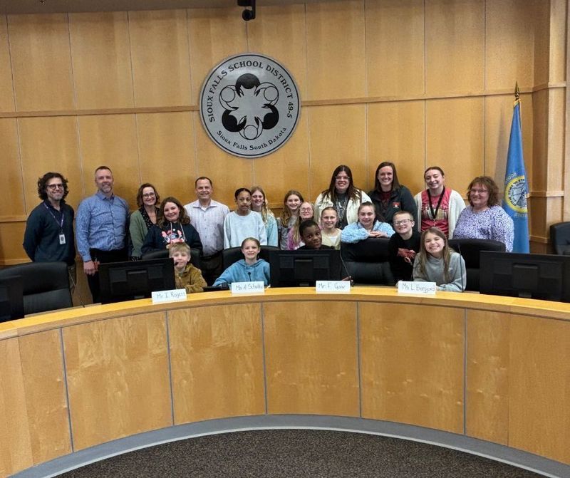 A group of students and District leaders stand in the IPC board room behind the school board desk smiling