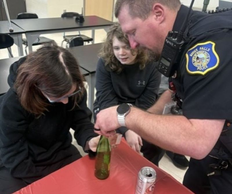 two students stand next to a police officer who is demonstrating how to capture a fingerprint from a pop bottle
