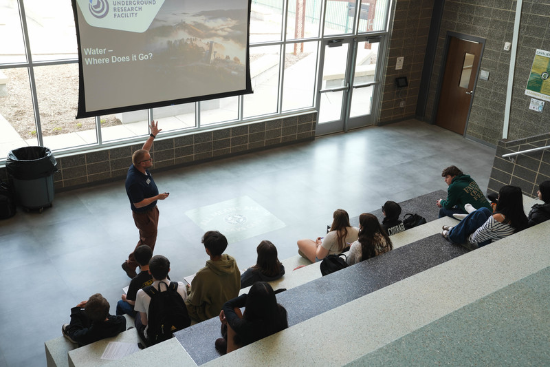 A man gives a presentation to a group of high school students in front of a display that says "Where does water go?"