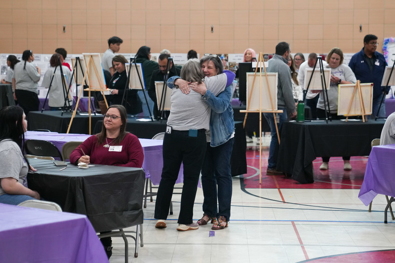 Two women hug in a gymnasium, behind them are pieces of art on easels, but only the backs of the canvases are visible. People can be seen looking at said art. 