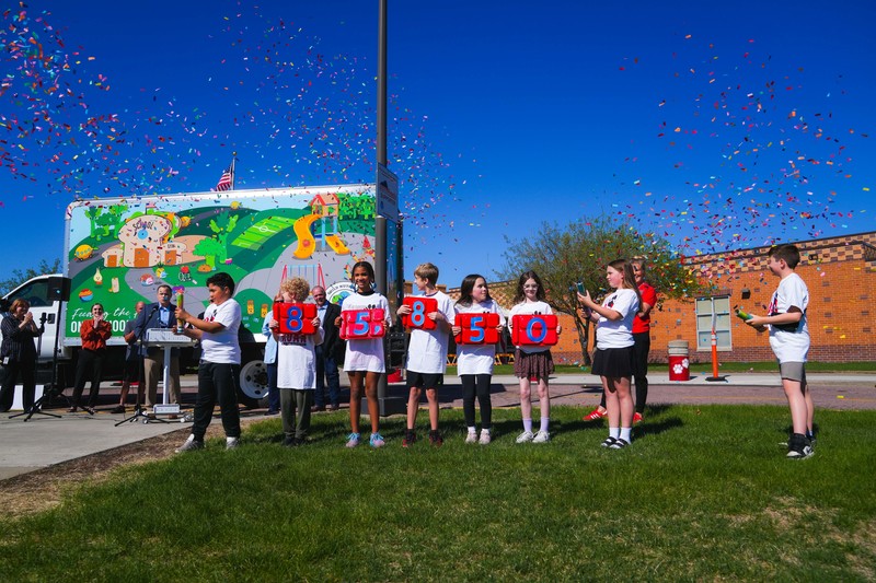 Confetti flies in the air in front of a school with a lunch truck in front of it. Young students hold up lunch trays with the number 85,850  on them. 