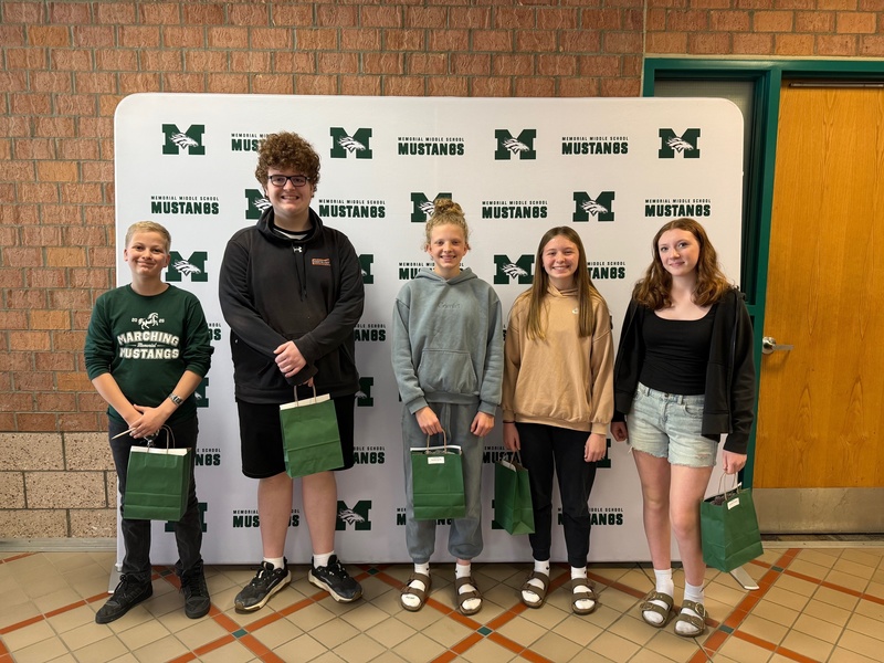 Five 8th grade students stand smiling in front of the Memorial Middle School Mustangs step-and-repeat backdrop. Several students are holding green MMS gift bags. The group is dressed casually in school and athletic wear.