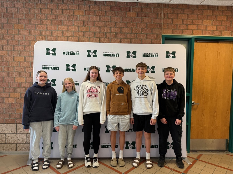 Six 7th grade students stand smiling in front of the Memorial Middle School Mustangs step-and-repeat backdrop. The group is dressed casually in hoodies and athletic wear.
