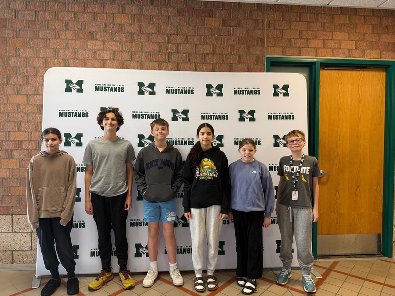 Six 6th grade students stand smiling in front of the Memorial Middle School Mustangs step-and-repeat backdrop. The group is dressed casually in a variety of hoodies and athletic wear.
