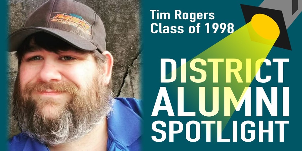 District Alumni Spotlight graphic featuring a close-up photo of Tim Rogers, Class of 1998. He is smiling, has a full beard, and is wearing a brown cap and blue shirt. The right side of the graphic shows a spotlight illustration shining onto the words “District Alumni Spotlight,” with his name and graduation year displayed at the top.