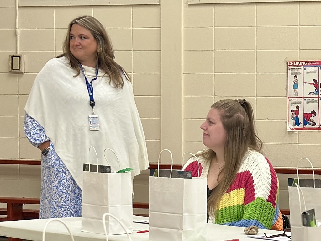 Two women are pictured in a classroom or meeting space. One woman, standing on the left, wears a white poncho over a blue patterned dress and has a lanyard with an ID badge around her neck. She is looking to the side with her hands in her pockets. The woman on the right, seated at a table, is smiling and wearing a colorful rainbow-striped crocheted sweater. On the table in front of them are white gift bags and papers. A “Choking” safety poster is visible on the wall in the background.