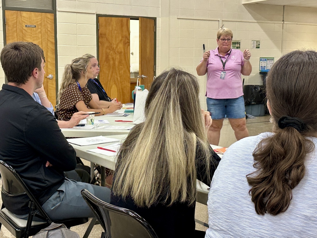 A group of adults sit at tables listening to a woman standing at the front of the room giving a presentation. The woman, wearing a pink polo shirt and denim shorts with a lanyard around her neck, gestures with both hands while speaking. Participants are seated with papers, pens, and bags on the tables in front of them, paying attention to the presenter. The setting appears to be a classroom or meeting room with beige walls and wooden doors in the background.