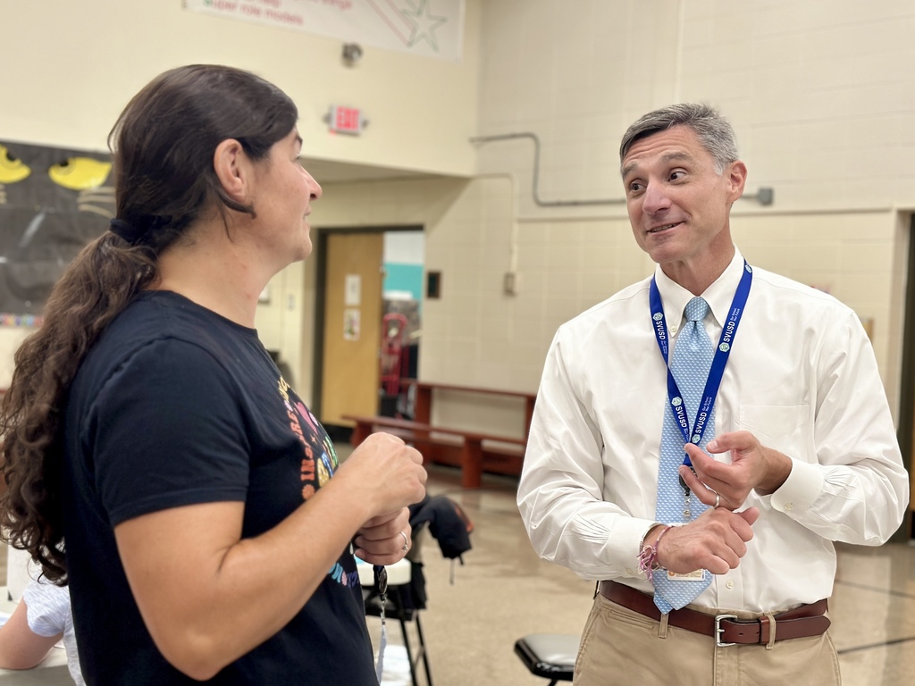 Two adults are engaged in conversation inside a school cafeteria. The person on the left, with long dark hair in a ponytail and wearing a black T-shirt, is listening attentively. The person on the right, wearing a white dress shirt, light blue tie, and khaki pants, is speaking while gesturing with his hands. He has a blue lanyard around his neck. The background shows a beige wall, door, and classroom furnishings.