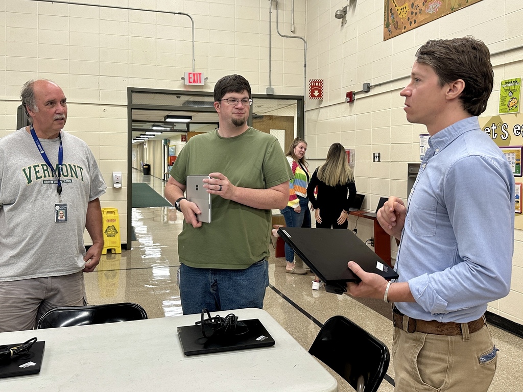 Three adults are standing and talking in a school hallway near a table. The person on the left is wearing a gray Vermont T-shirt and lanyard, the person in the middle is holding a tablet and wearing a green shirt, and the person on the right is holding a closed laptop and wearing a light blue button-down shirt. In the background, two women are conversing near a laptop; one is wearing a colorful striped sweater. The setting includes cream-colored walls, posters, and a hallway extending behind them.