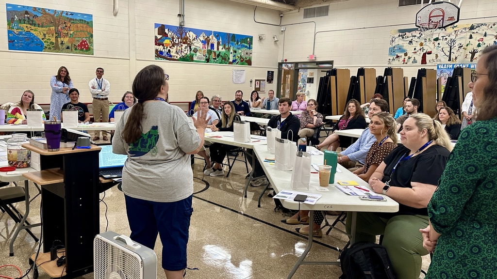 A large group of adults sits at tables arranged in a U-shape inside a school gymnasium for a meeting or training session. A woman wearing a Slate Valley T-shirt stands at the front, speaking to the group. Attendees are listening attentively, with papers, pens, and white gift bags in front of them on the tables. Some participants are seated in folding chairs, while a few others stand in the back near colorful murals painted on the walls. A basketball hoop and stacked cafeteria tables are visible in the background, indicating a multipurpose school space.