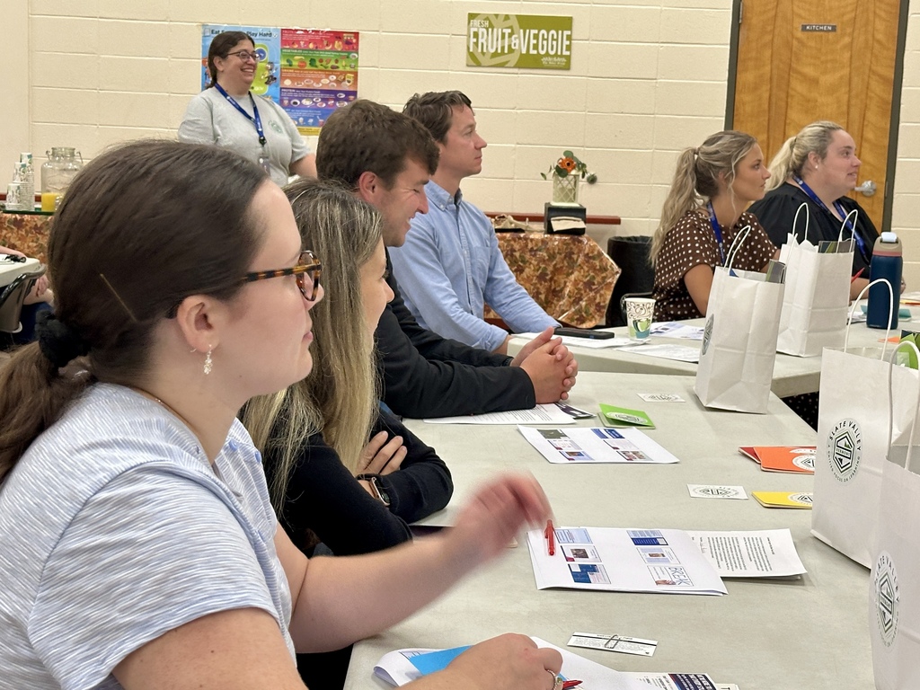 Several adults sit at tables in a classroom or meeting space, attentively facing forward during a session. They have papers, pens, water bottles, and white bags with the Slate Valley logo on the table in front of them. A woman in glasses and a ponytail is in the foreground, holding a red pen. Other participants are seated in a row, some leaning forward with hands clasped. In the background, a person wearing a Slate Valley shirt and lanyard stands and smiles near a refreshment table. Posters and signs decorate the walls.