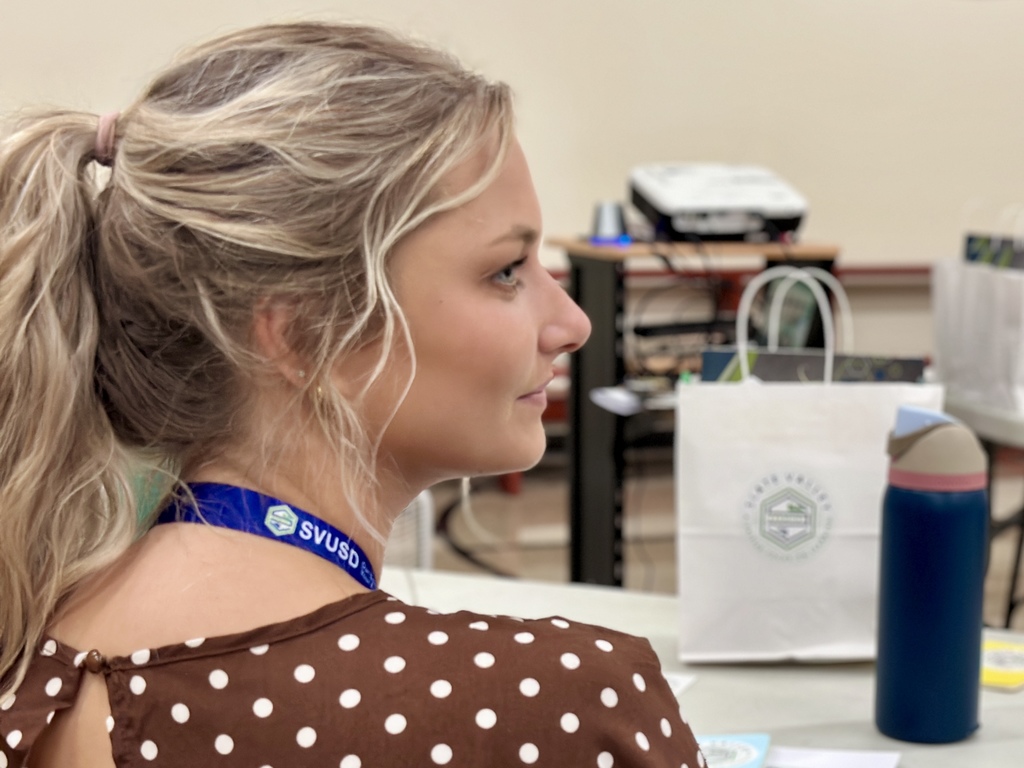 A close-up of a young woman with blonde hair tied in a ponytail, wearing a brown polka-dot top and an SVUSD lanyard. She is seated at a table with a white gift bag featuring the Slate Valley logo and a reusable water bottle in front of her. In the background, other bags and a projector are visible on a table, indicating a classroom or meeting setting.