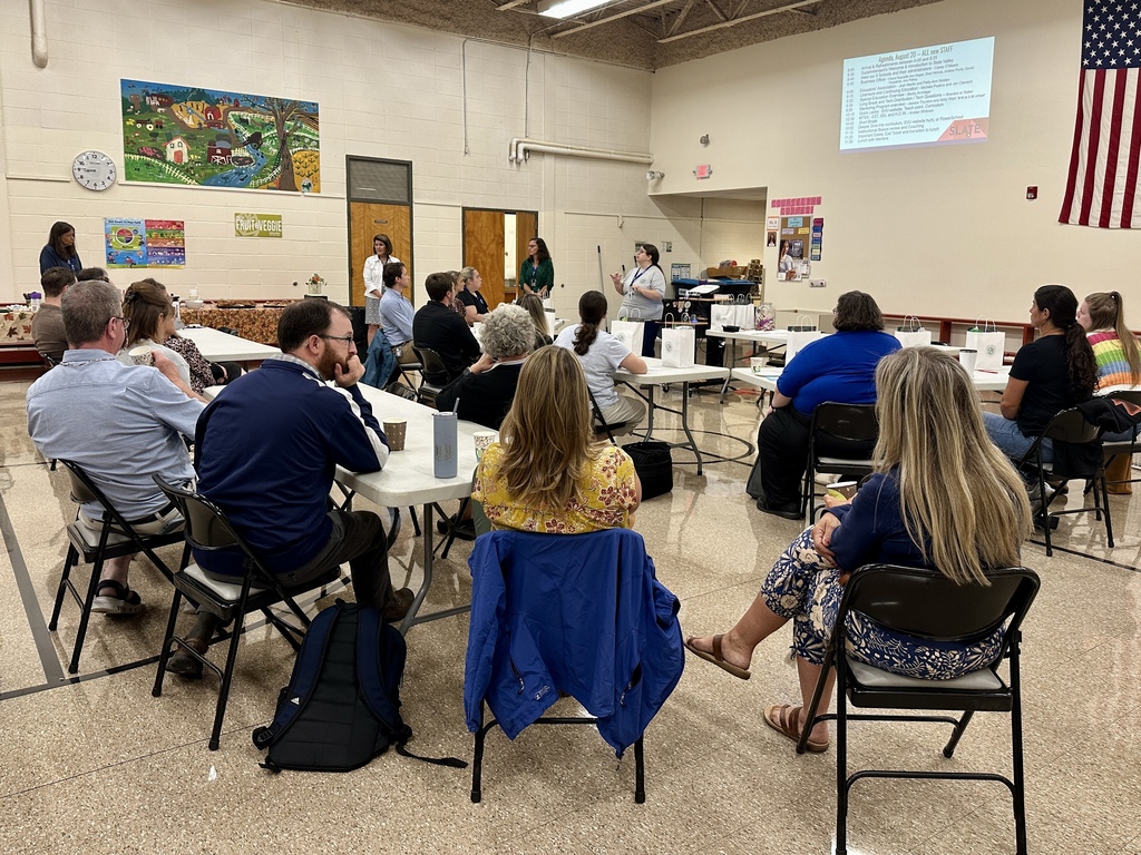 A group of adults is seated in a school gymnasium, attending a meeting or training session. Tables are arranged in a U-shape, with papers, drinks, and white gift bags placed in front of participants. At the front of the room, a woman in a gray Slate Valley T-shirt speaks to the group while standing near a podium. A large agenda is projected onto the wall behind her, and an American flag hangs nearby. Colorful murals decorate the walls, and attendees are listening attentively.