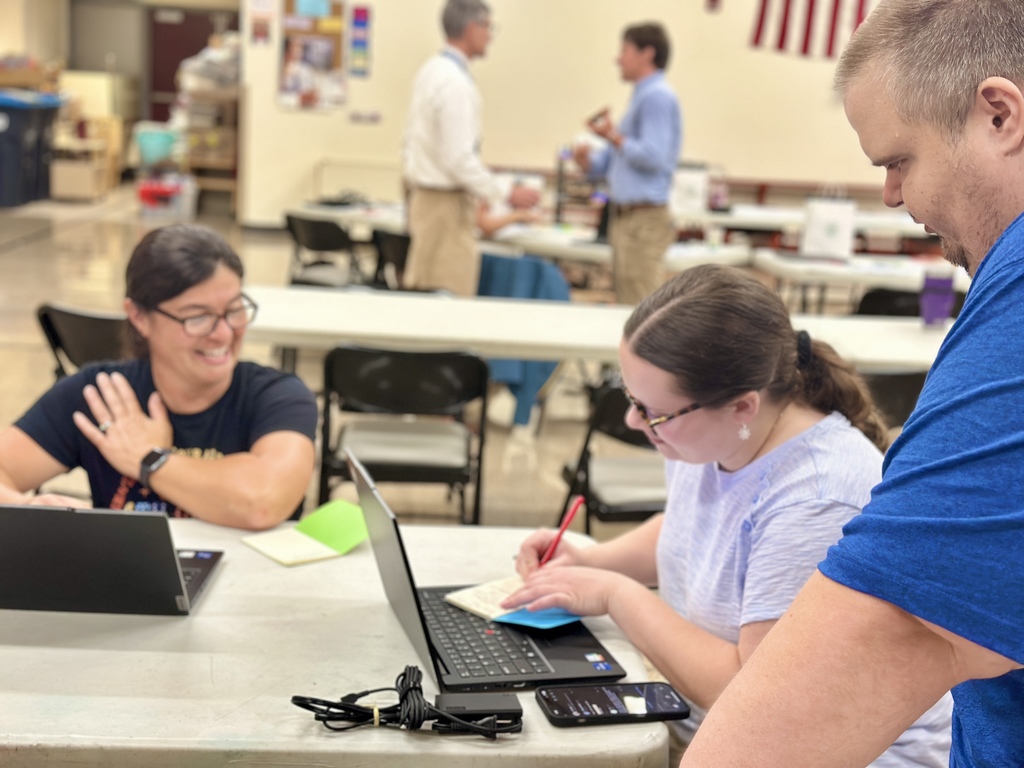 Three adults are gathered around a table working collaboratively. A woman on the left is smiling while seated at a laptop. In the center, another woman is writing on a sticky note placed on her laptop, with a phone and charger on the table in front of her. A man on the right leans over to observe. The background shows additional people conversing near tables, with an American flag visible on the wall. The setting is in a school cafeteria..