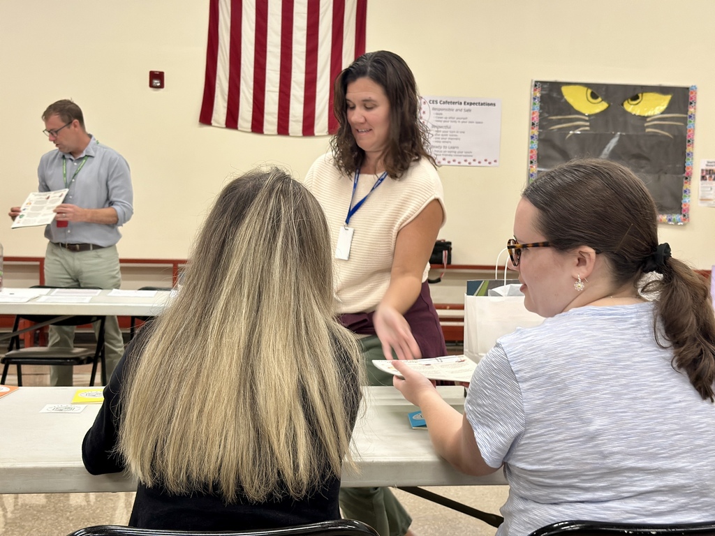 A woman in a cream-colored top and lanyard hands a paper to another woman seated at a table, who is wearing glasses and a light blue shirt. Another person with long blonde hair sits beside her, facing forward. In the background, a man in a light blue shirt looks down at a paper while standing at a separate table. The room has school posters on the walls, including a large black poster with yellow eyes, and an American flag is displayed. White bags and papers are spread across the tables where participants are seated.