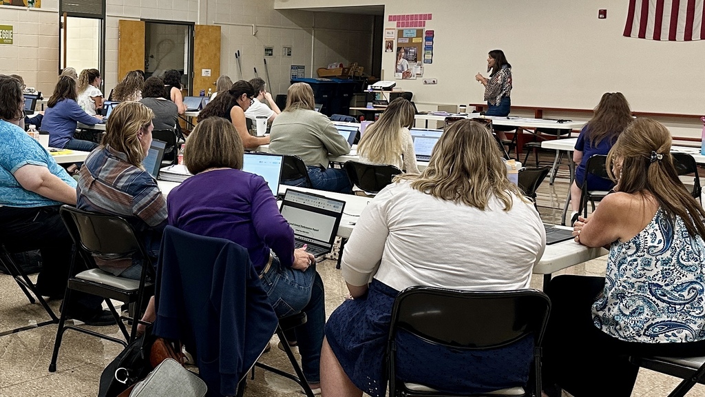 Educators sit at tables with laptops, listening to a presenter speaking at the front of a school cafeteria.