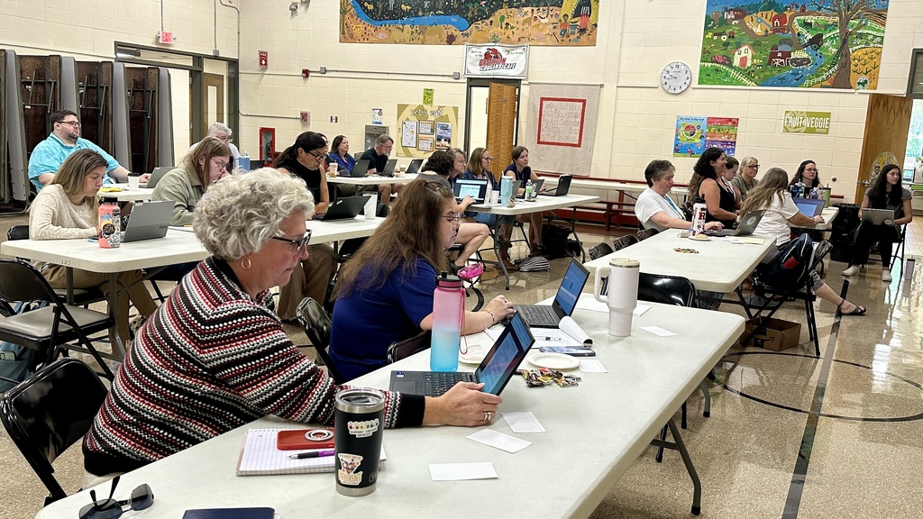 Educators sit at tables with laptops, listening to a presenter speaking at the front of a school cafeteria.