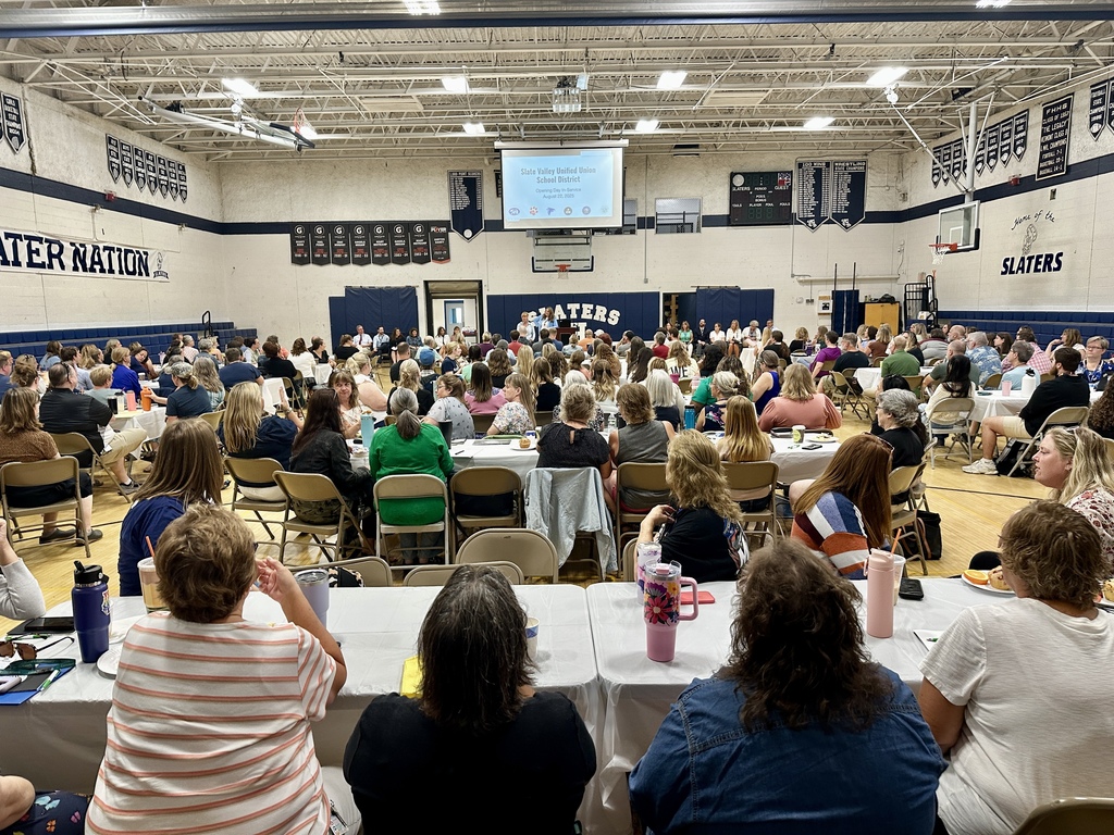 A large group of teachers and staff sit at tables in a school gymnasium during a district-wide meeting, facing a presentation screen at the front.